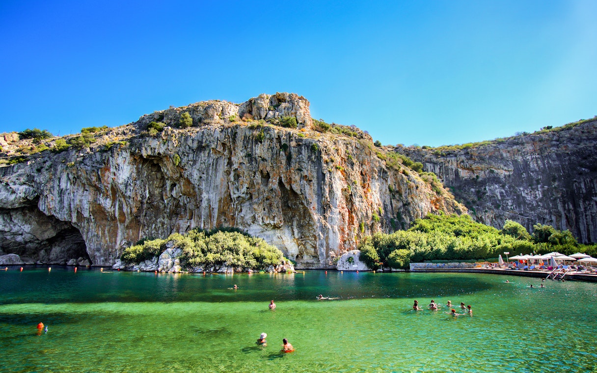 Swimmers enjoying Vouliagmeni Lake near Athens with rocky cliffs and greenery.