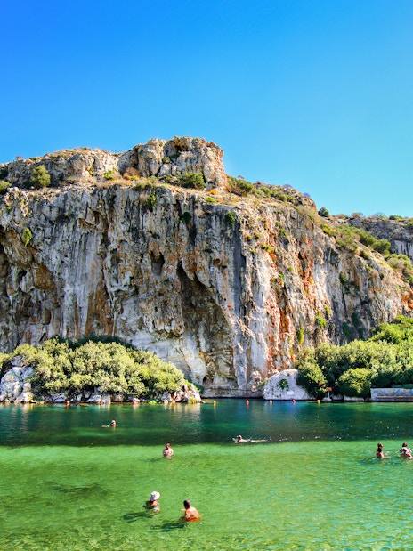 Swimmers enjoying Vouliagmeni Lake near Athens with rocky cliffs and greenery.