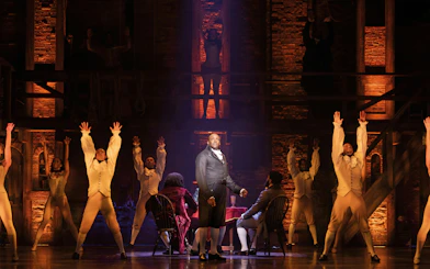Hamilton cast performing on West End stage with actors in period costumes.