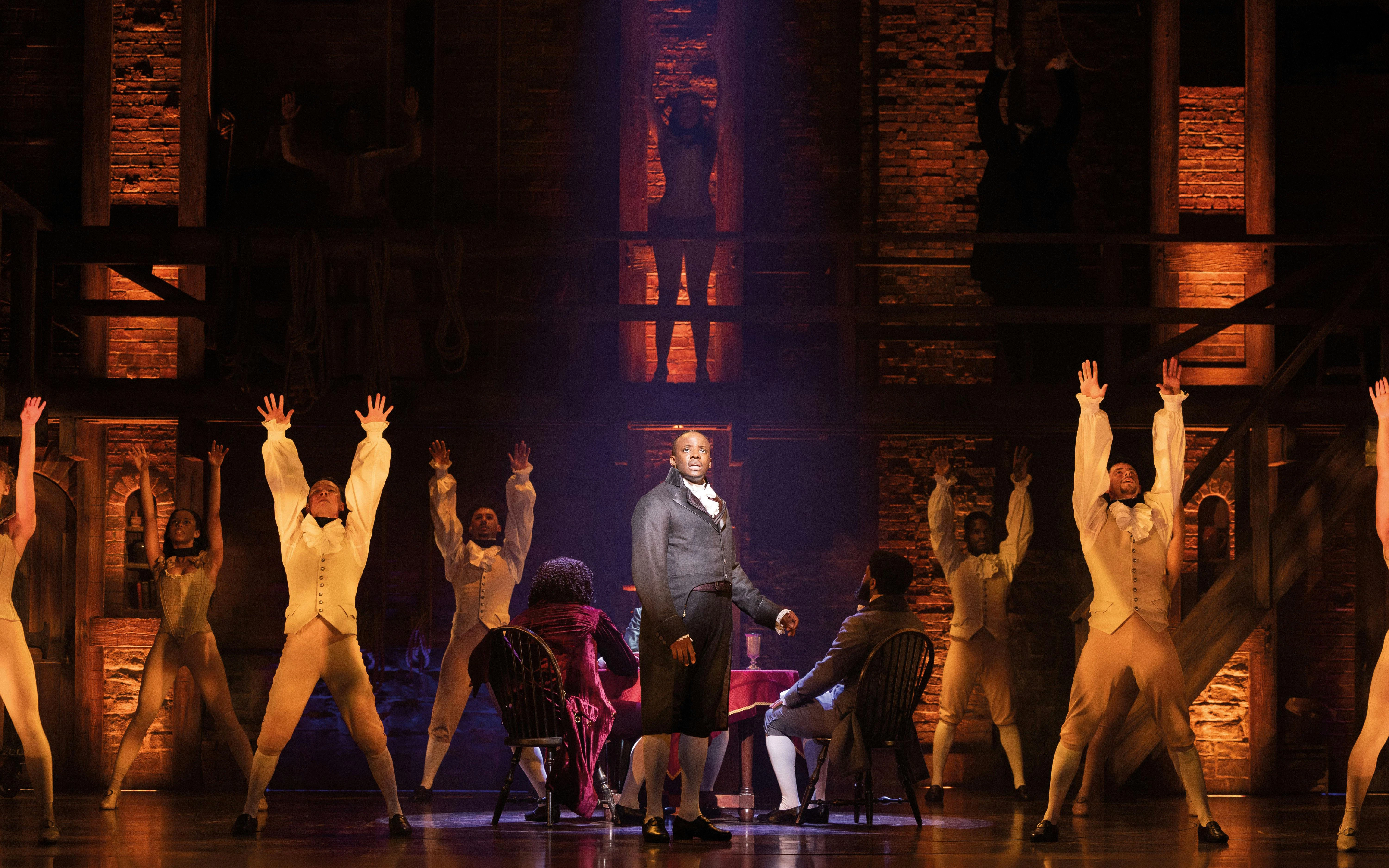 Hamilton cast performing on West End stage with actors in period costumes.