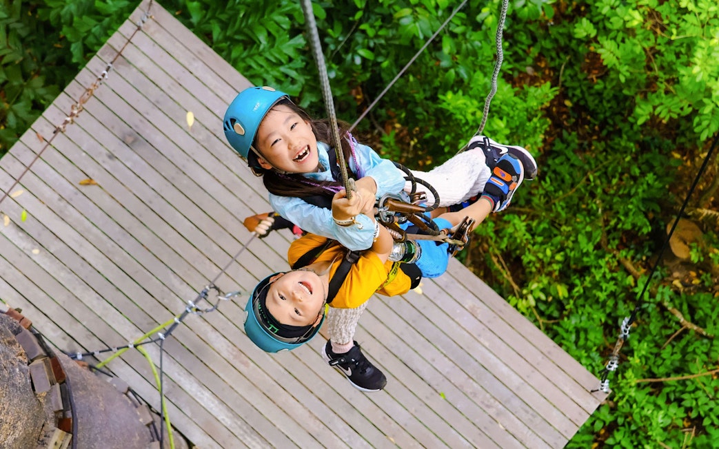 Children ziplining at Flying Hanuman, Phuket, on a wooden platform amidst lush greenery.