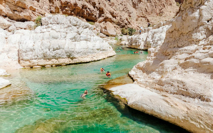 Tourists swimming in the turquoise waters of Wadi Shab, Oman, surrounded by rocky cliffs.