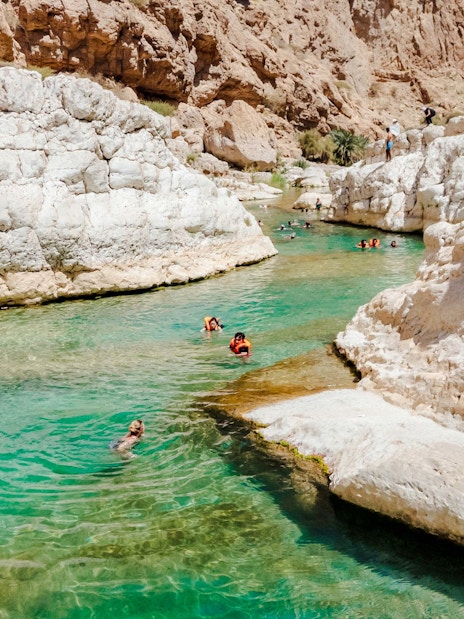Tourists swimming in the turquoise waters of Wadi Shab, Oman, surrounded by rocky cliffs.
