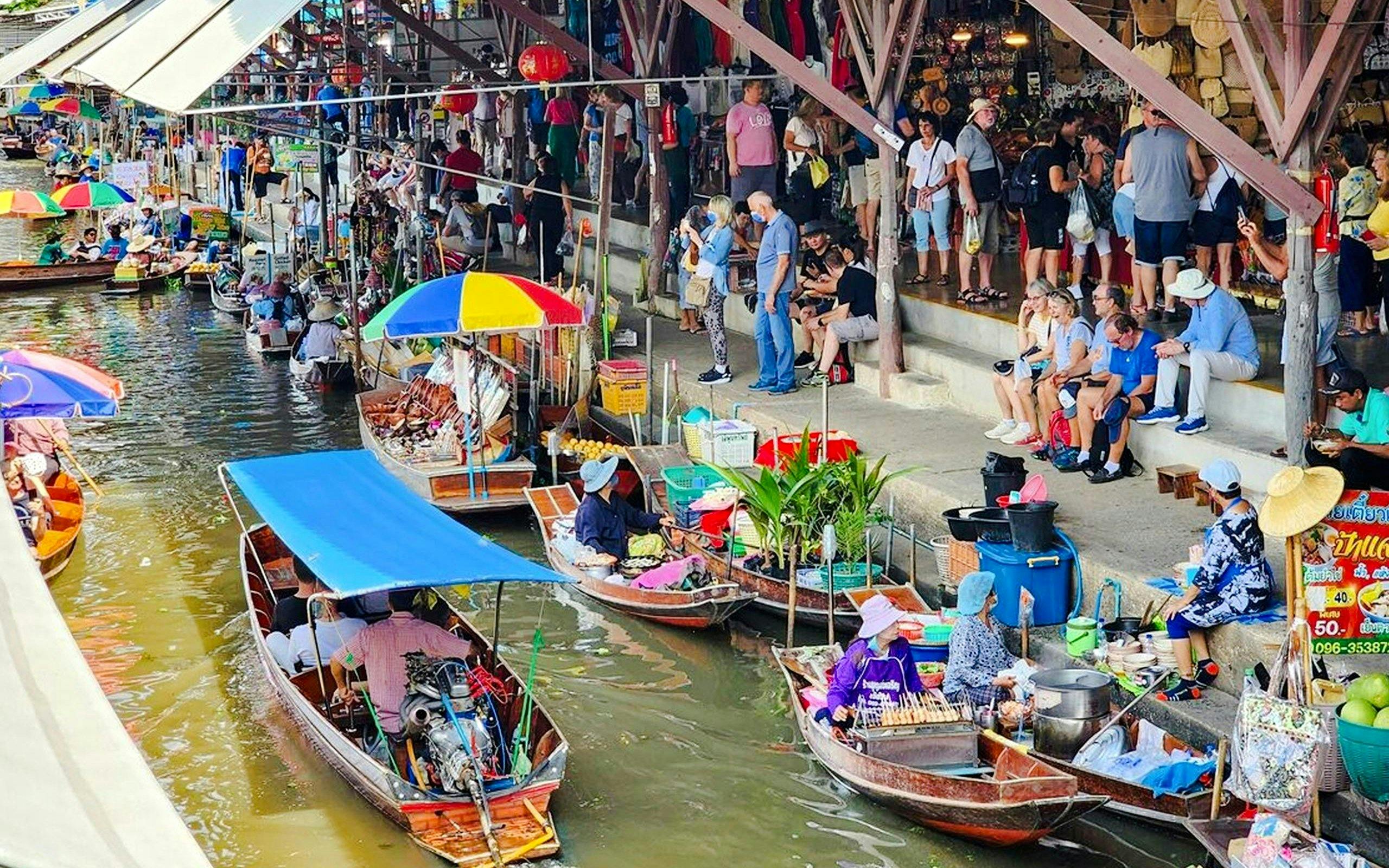 Boats with vendors at Damnoen Saduak Floating Market, Thailand, with tourists on the canal bank.