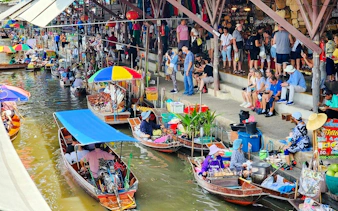Boats with vendors at Damnoen Saduak Floating Market, Thailand, with tourists on the canal bank.