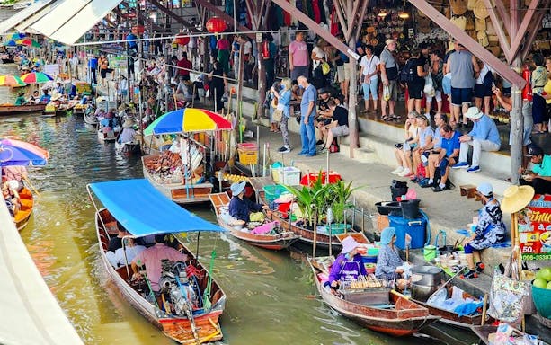 Boats with vendors at Damnoen Saduak Floating Market, Thailand, with tourists on the canal bank.