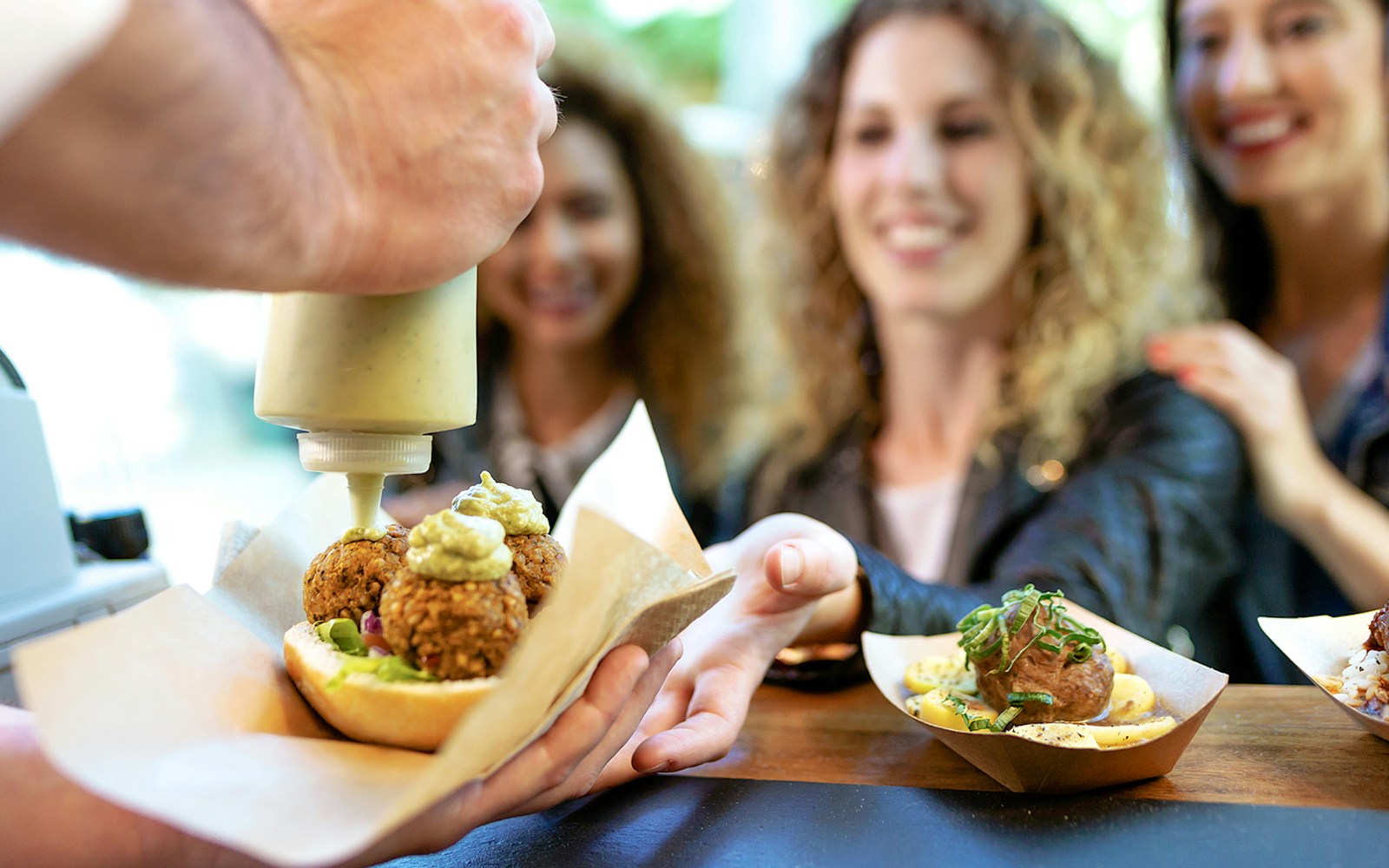 Women receiving food at a market stall, with falafel and sauce being served.