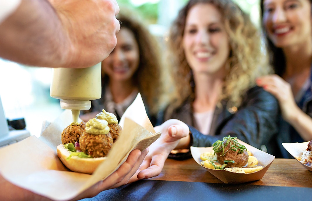 Women receiving food at a market stall, with falafel and sauce being served.