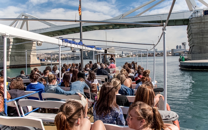 Passengers on Las Golondrinas boat trip under a bridge in Barcelona harbor.