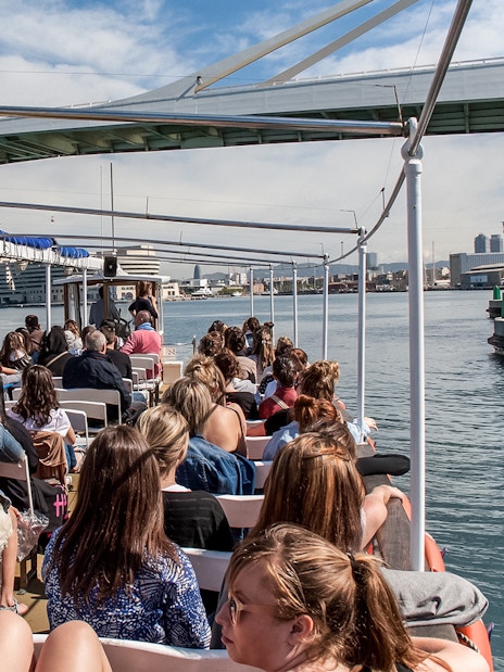 Passengers on Las Golondrinas boat trip under a bridge in Barcelona harbor.
