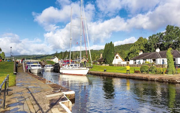 Boats docked along the canal in Fort Augustus, Scotland, with cottages and greenery nearby.