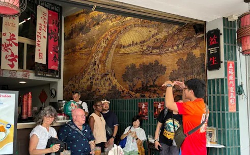 Tour group in Bangkok Chinatown café with traditional mural and guide explaining.