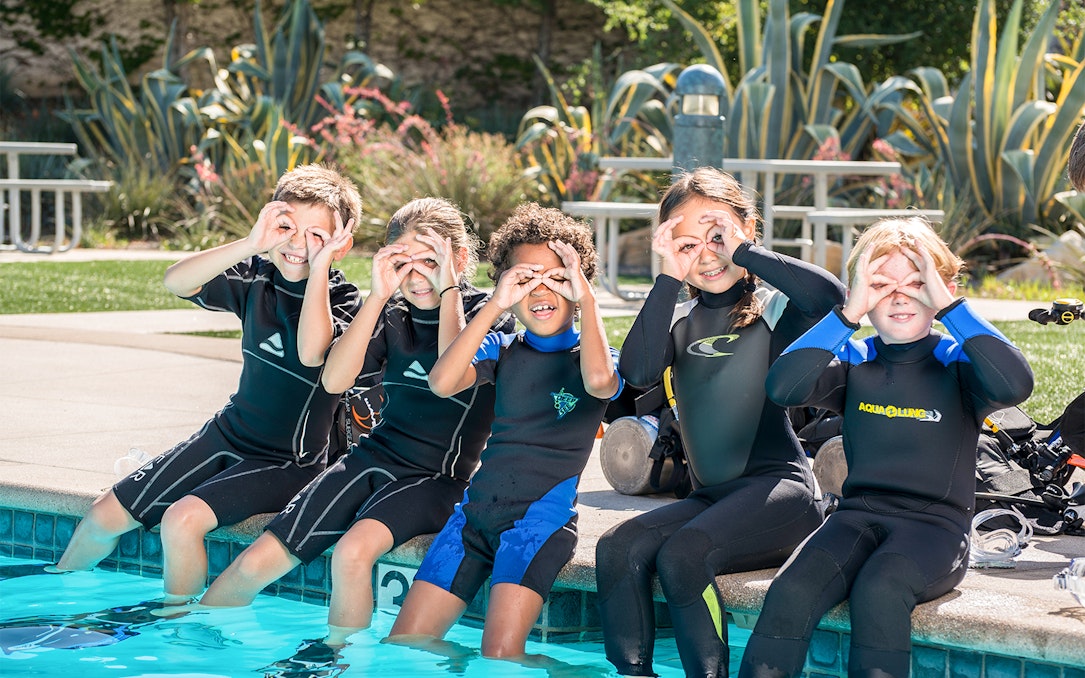 Children in wetsuits sitting by a pool, preparing for PADI Discover Scuba Diving at Palm Jumeirah Dubai.