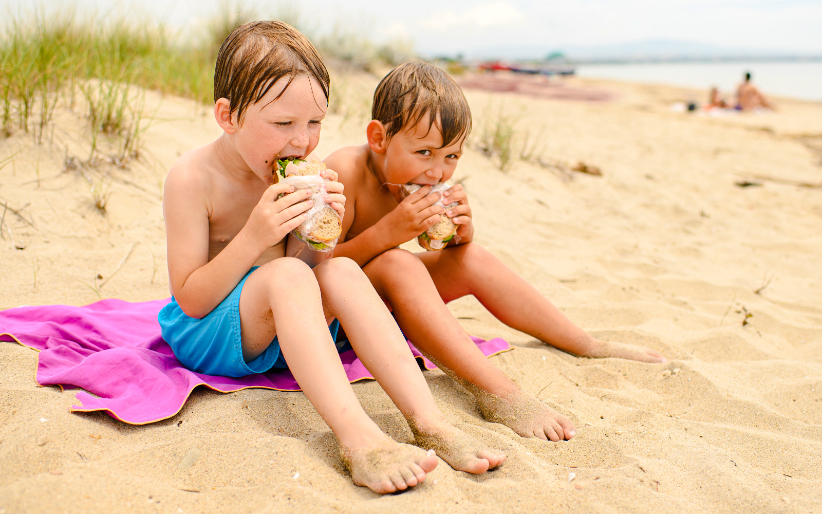 Boys enjoying sandwiches on a sandy beach.