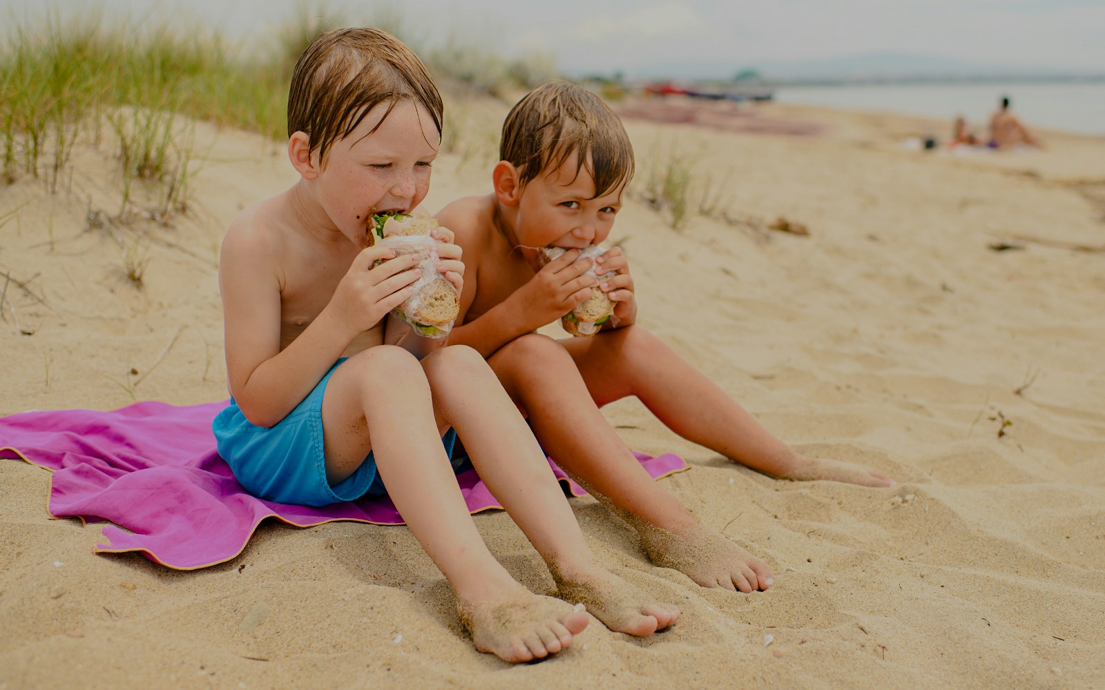 Boys enjoying sandwiches on a sandy beach.