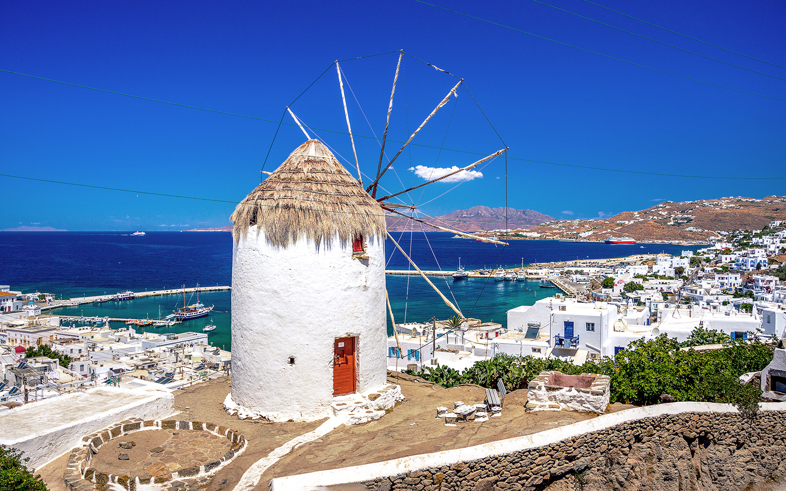Mykonos windmill overlooking the Aegean Sea with white buildings and harbor in view.