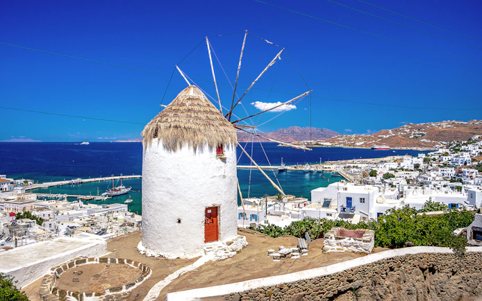 Mykonos windmill overlooking the Aegean Sea with white buildings and harbor in view.