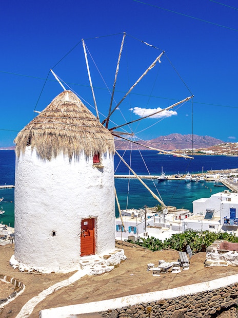 Mykonos windmill overlooking the Aegean Sea with white buildings and harbor in view.