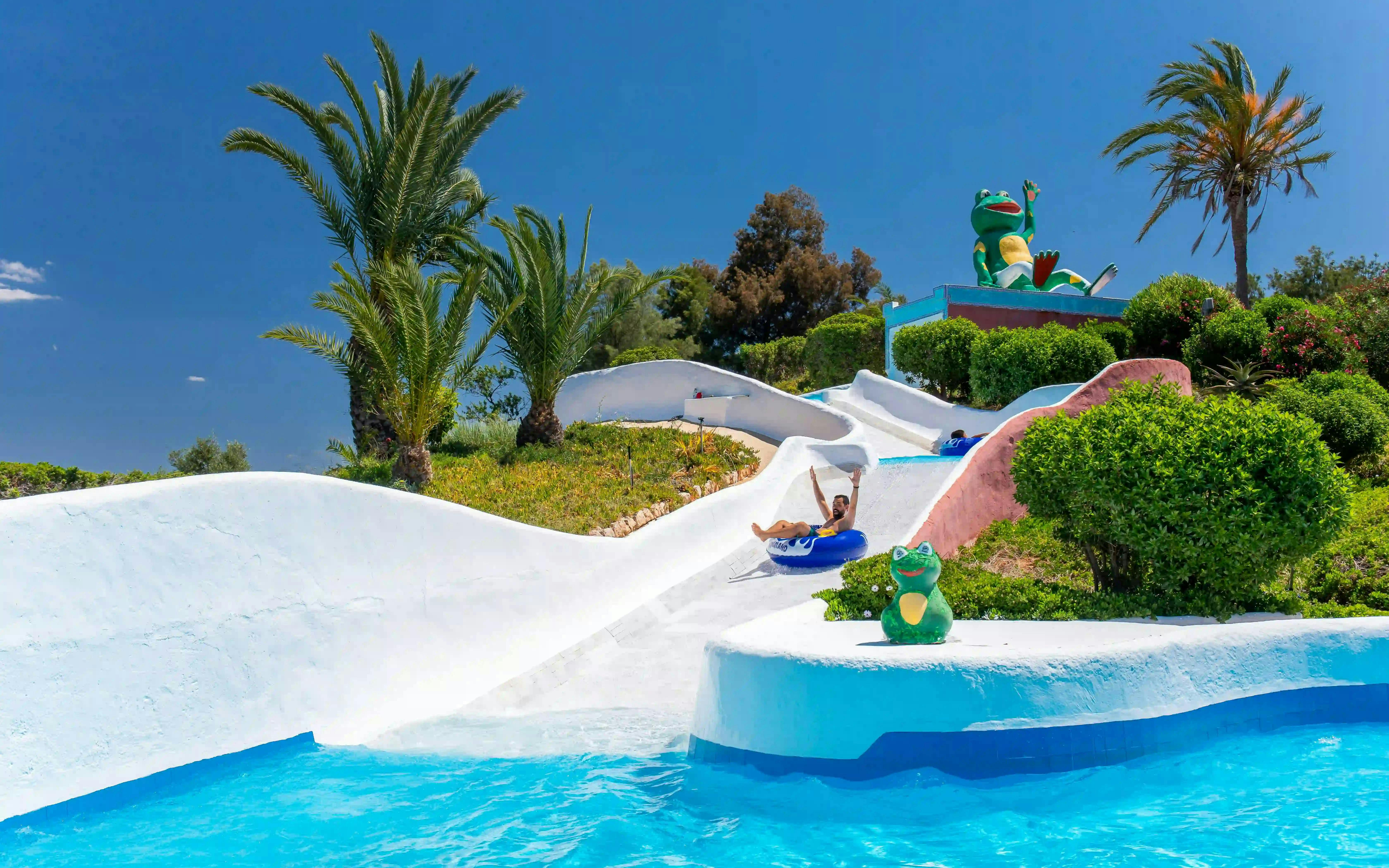 Visitors enjoying water rapids at Aqualand Algarve with palm trees and frog statue.