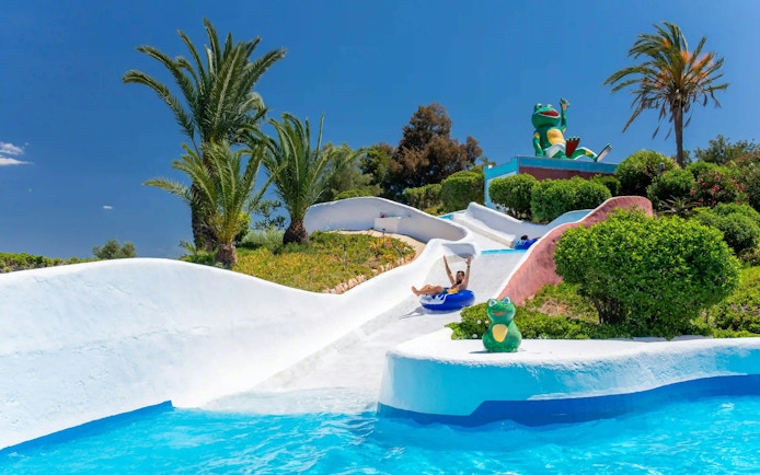 Visitors enjoying water rapids at Aqualand Algarve with palm trees and frog statue.