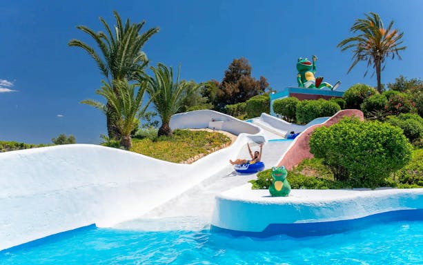 Visitors enjoying water rapids at Aqualand Algarve with palm trees and frog statue.