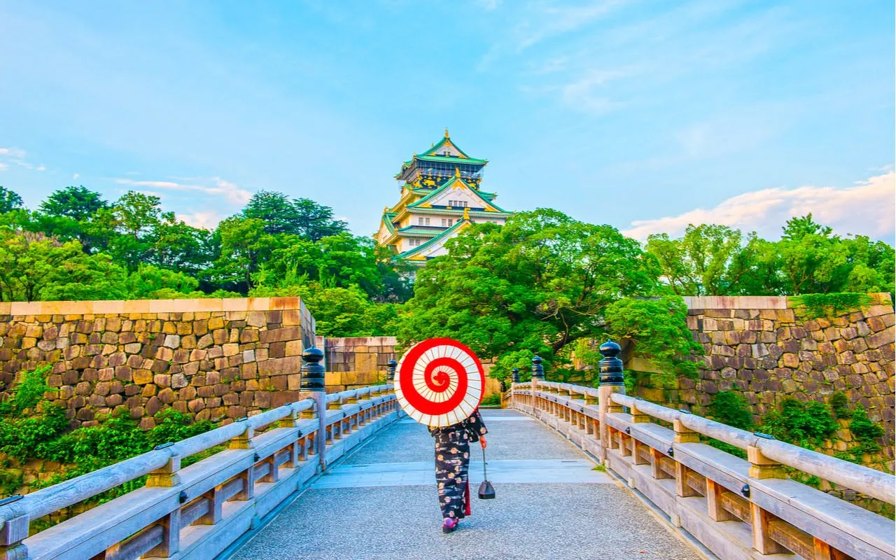 Person with parasol walking towards Osaka Castle on a stone bridge.