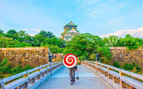 Person with parasol walking towards Osaka Castle on a stone bridge.