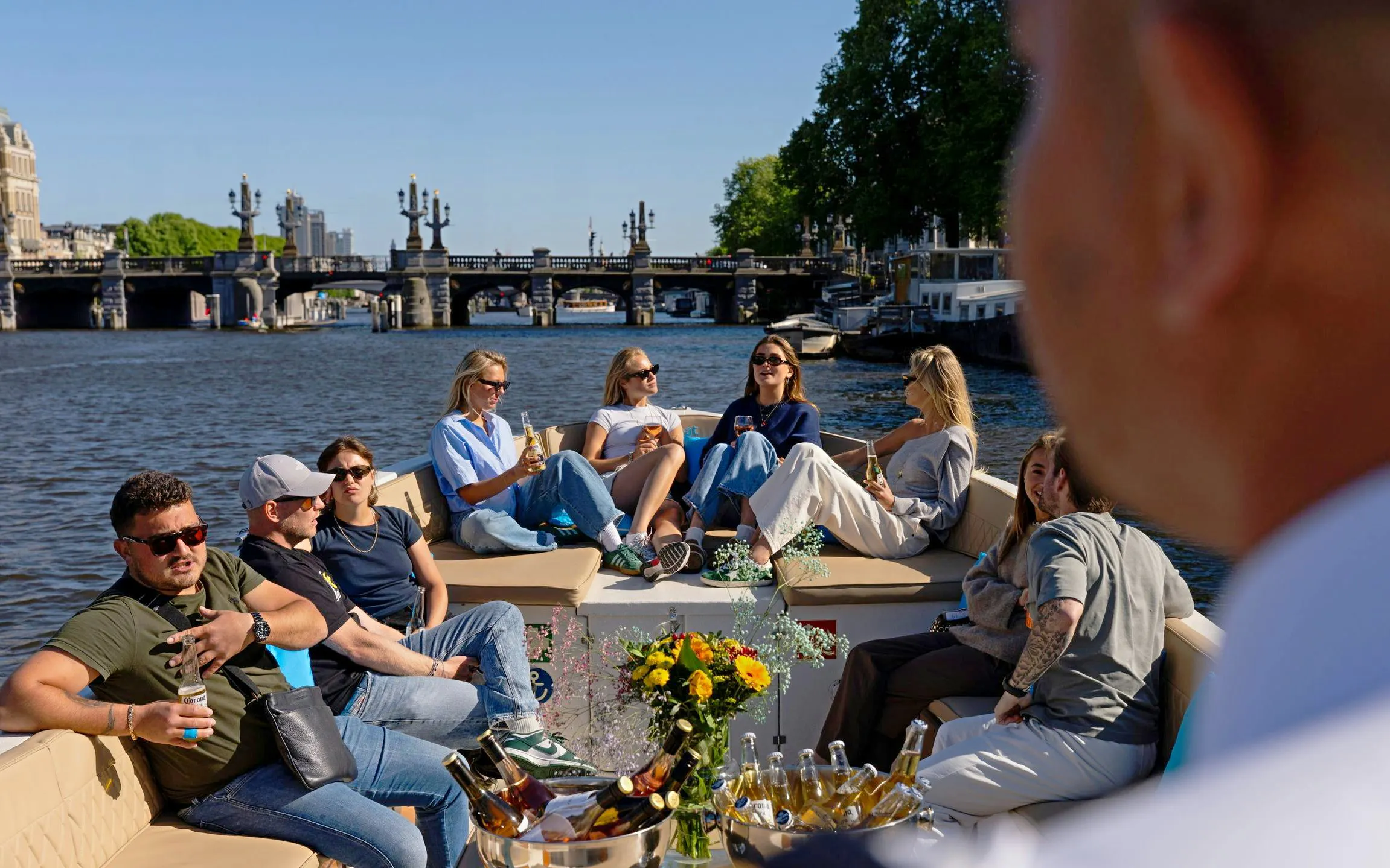 Group enjoying a canal cruise on an open electric boat in Amsterdam with a view of a historic bridge.