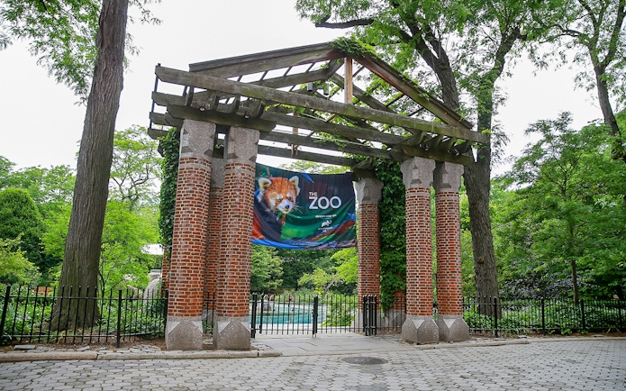 Entrance gate to a zoo with a banner featuring a red panda, surrounded by trees.