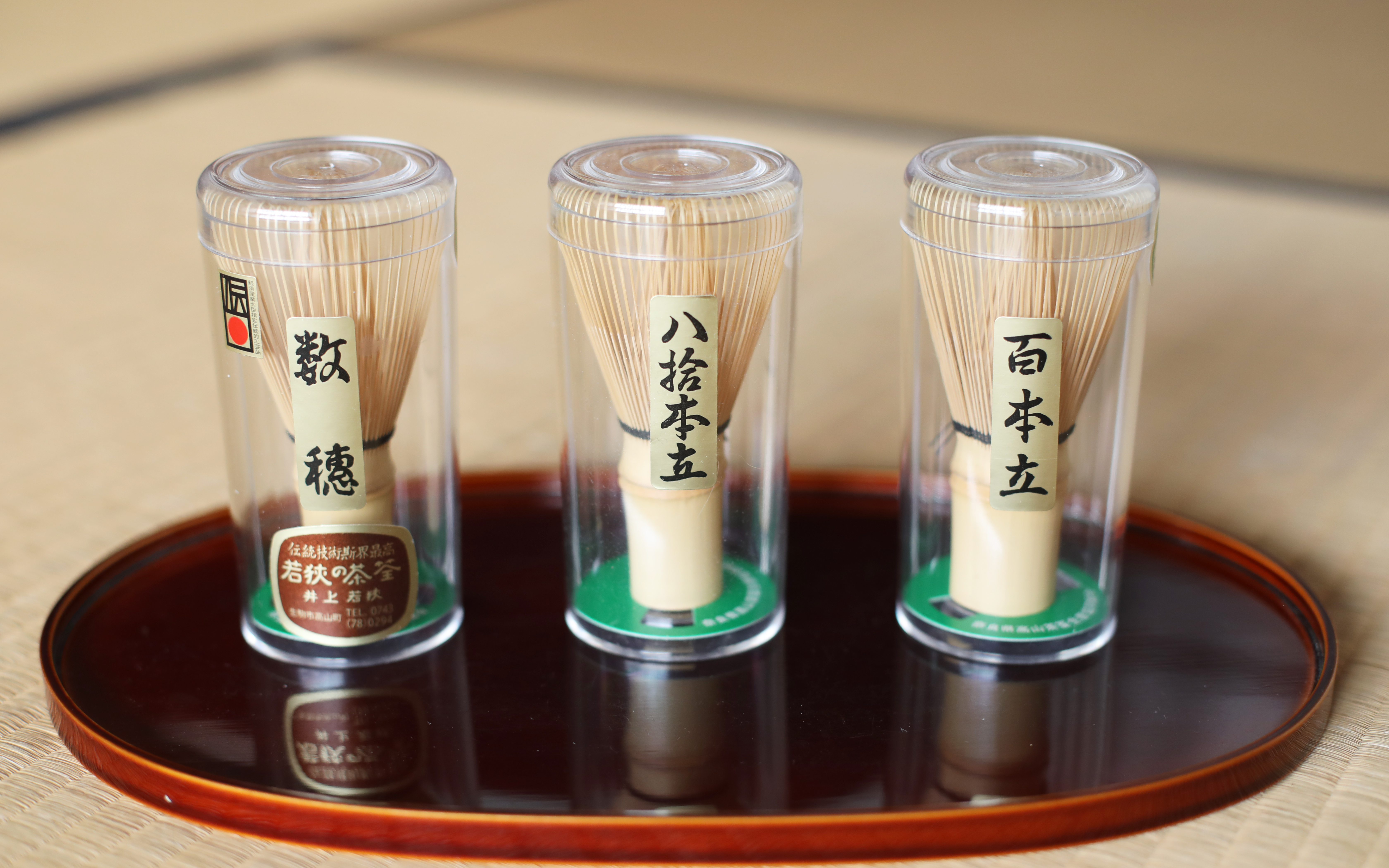 Bamboo matcha whisks in clear containers on a wooden tray.