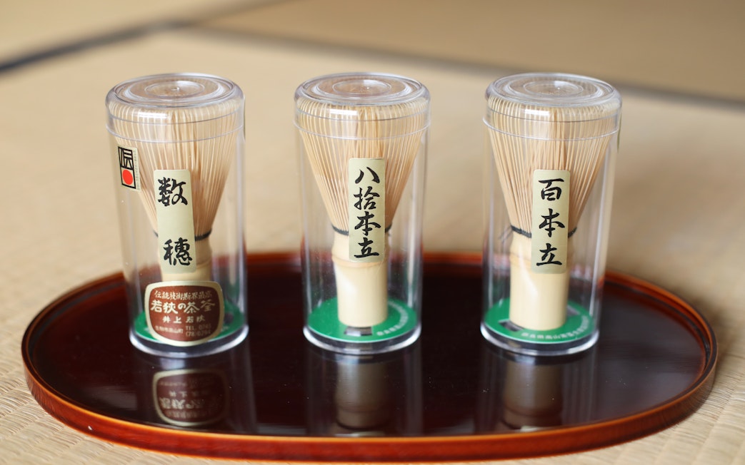 Bamboo matcha whisks in clear containers on a wooden tray.