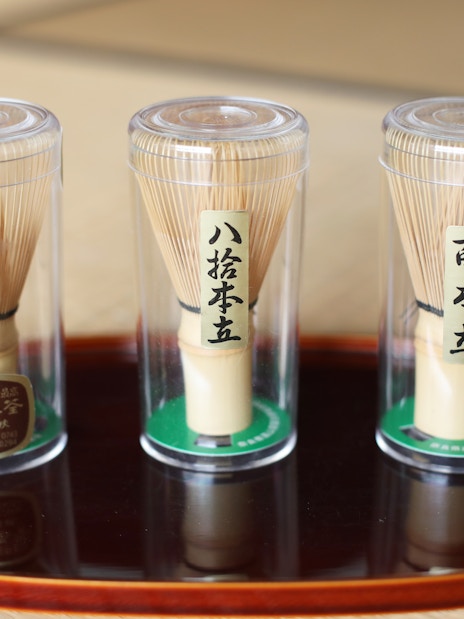 Bamboo matcha whisks in clear containers on a wooden tray.