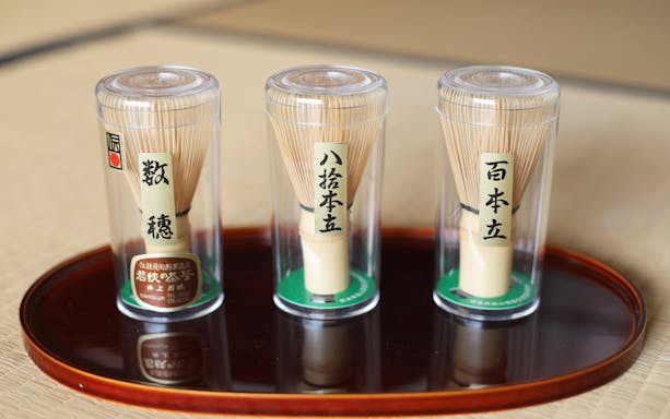 Bamboo matcha whisks in clear containers on a wooden tray.