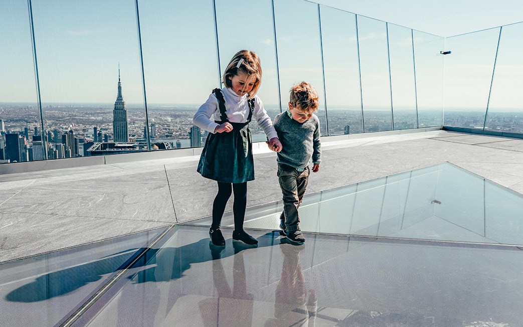Children walking on glass floor at Edge Observation Deck, New York City skyline in background.