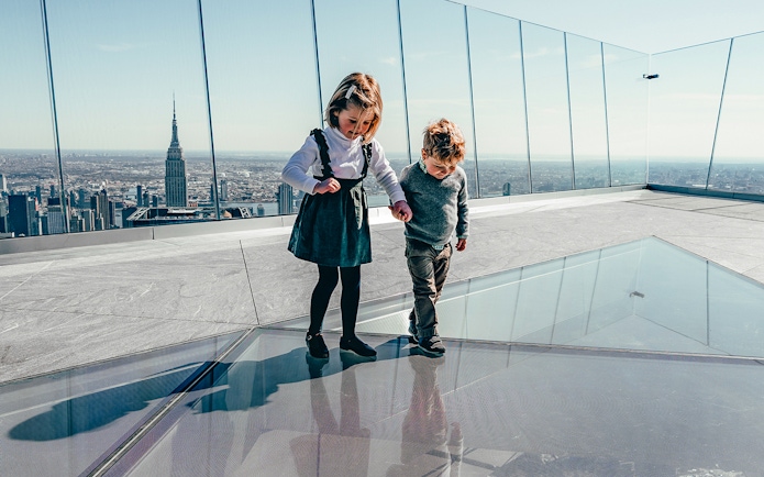 Children walking on glass floor at Edge Observation Deck, New York City skyline in background.