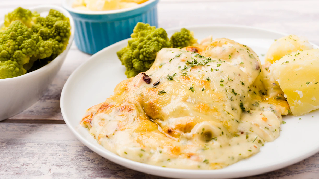 Cod fish casserole with cheese and seafood on a white plate, served with potatoes and romanesco cauliflower.
