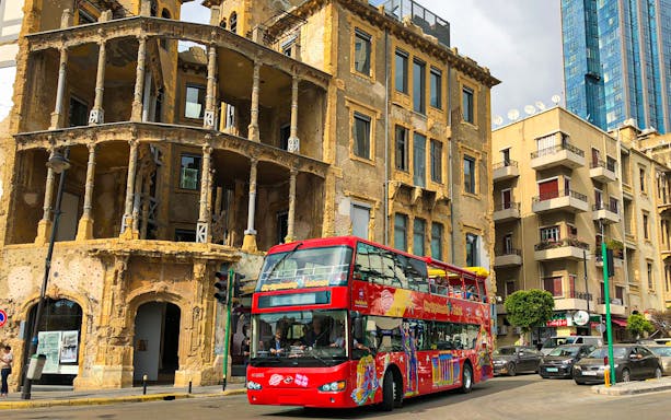 Red double-decker bus on Beirut street near historic building, part of Hop On Hop Off tour.