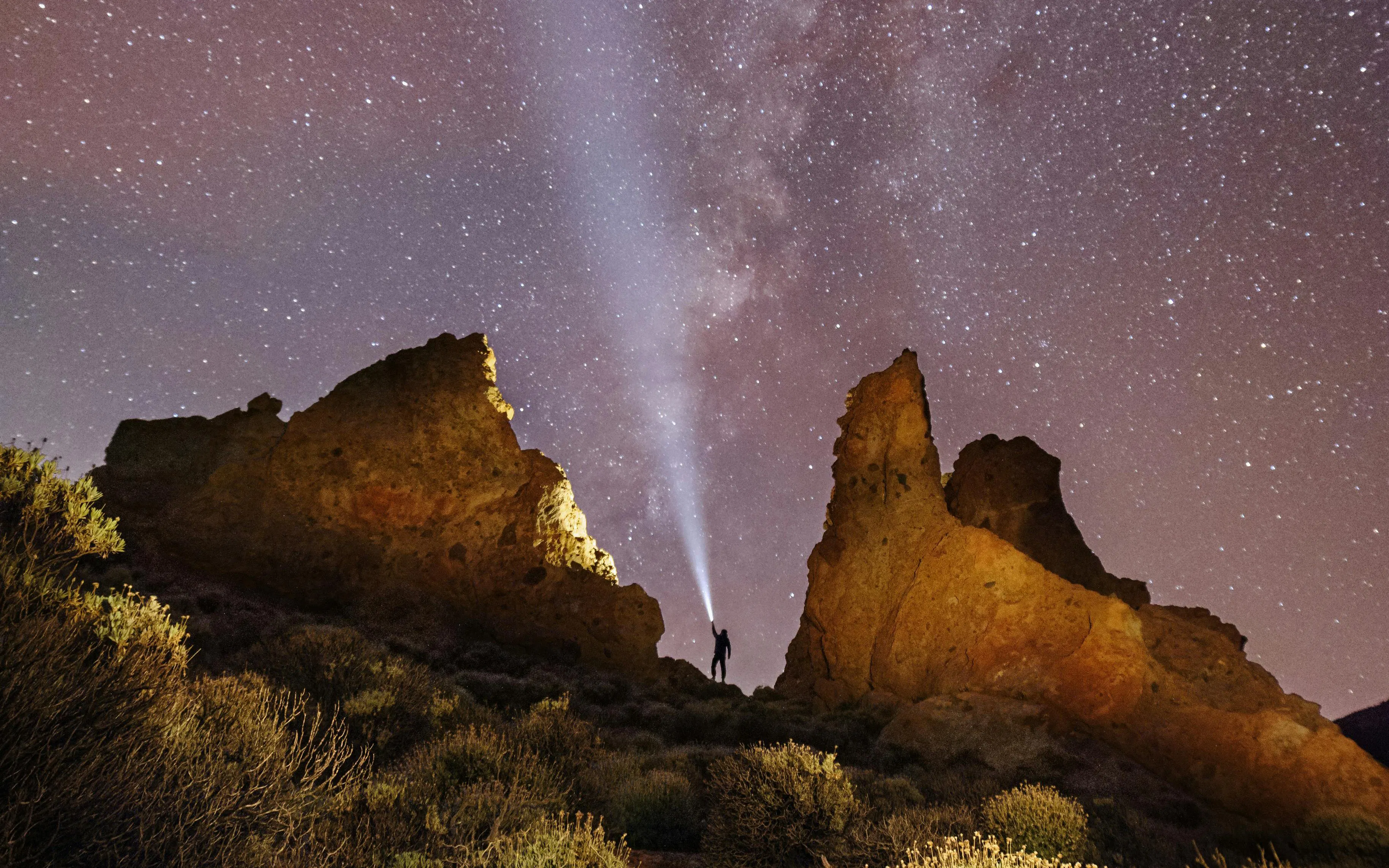 Person with flashlight near rock formations under Milky Way sky.