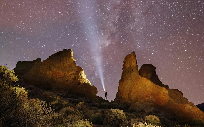 Person with flashlight near rock formations under Milky Way sky.