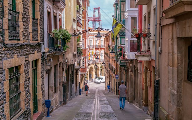 Narrow street in Bilbao with colorful buildings and the Vizcaya Bridge in the background.