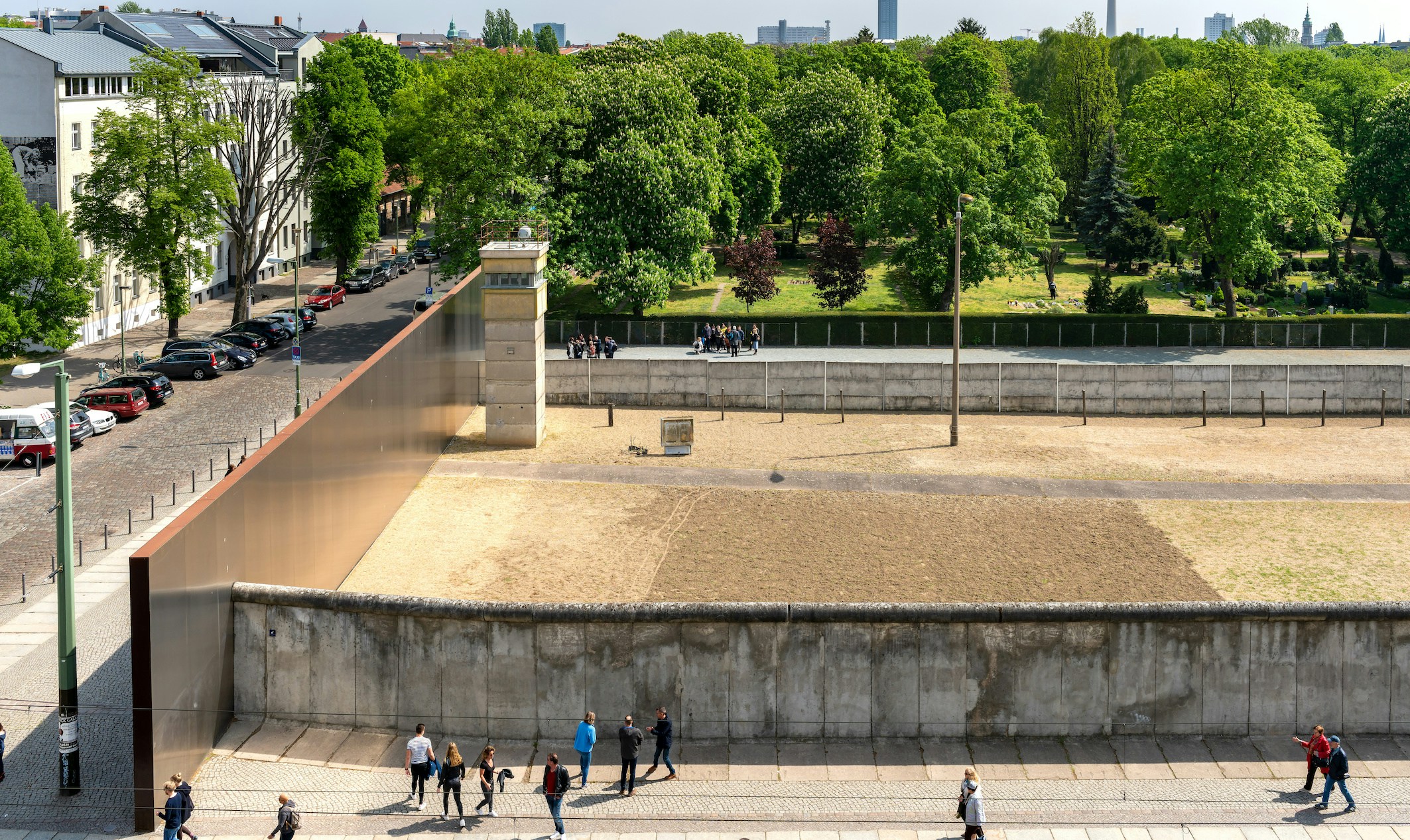 Gedenkstätte Berliner Mauer mit Grenzanlage, Wachturm und Besuchern, umgeben von Bäumen und Wohngebäuden.