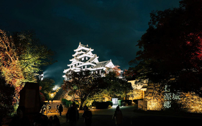 Okayama Castle illuminated at night with visitors walking nearby, Japan.