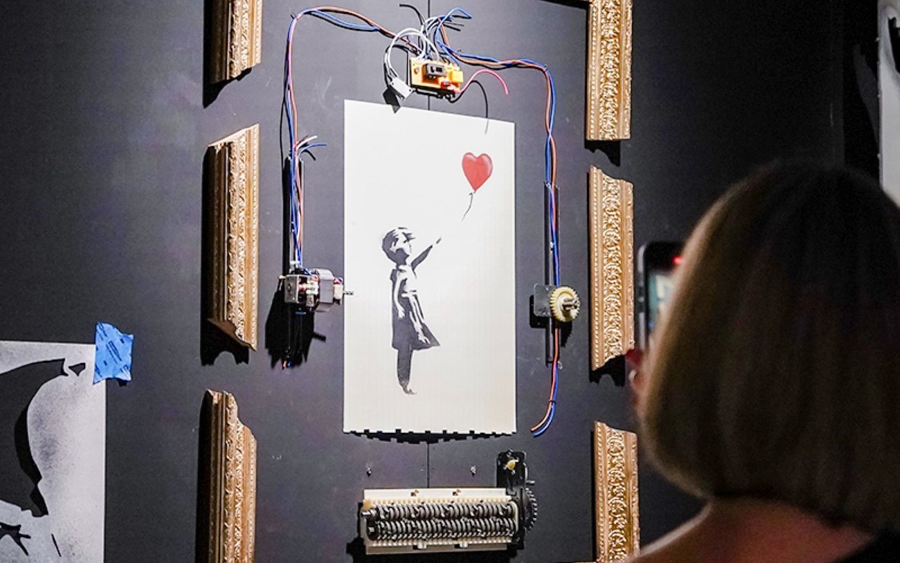 Banksy artwork with girl and balloon, partially shredded, displayed in a frame at Banksy Limitless exhibit.