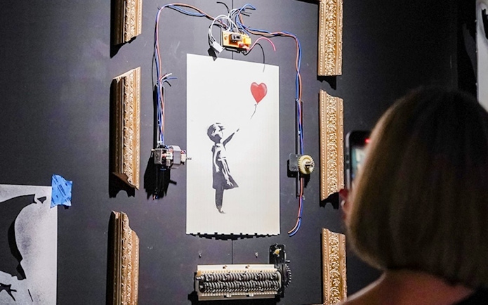 Banksy artwork with girl and balloon, partially shredded, displayed in a frame at Banksy Limitless exhibit.