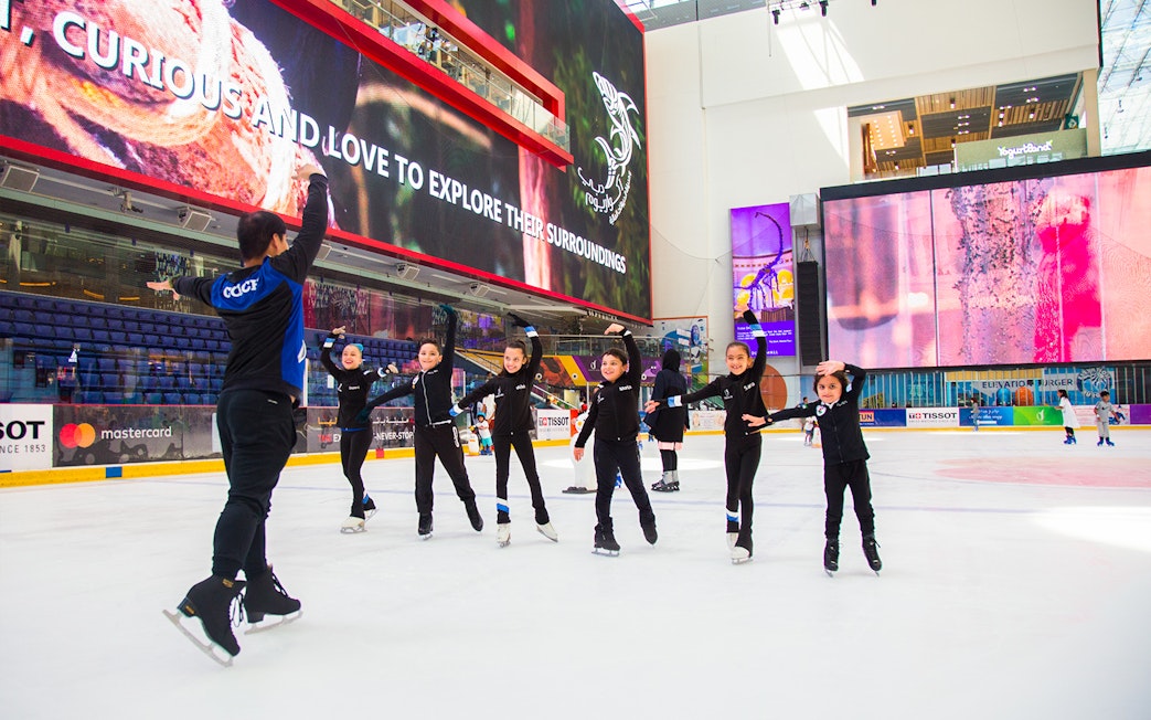 Children ice skating at Dubai Ice Rink under large digital screens.