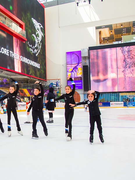 Children ice skating at Dubai Ice Rink under large digital screens.