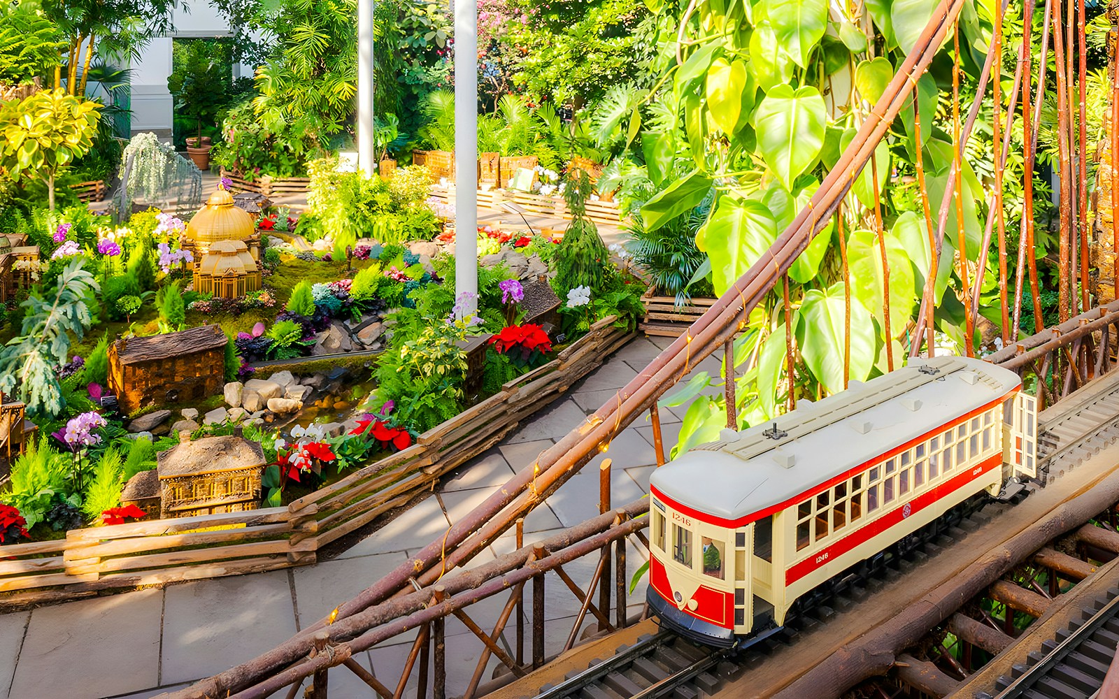 Miniature train on a bridge at NYBG Holiday Train Show, surrounded by lush greenery and model landmarks.