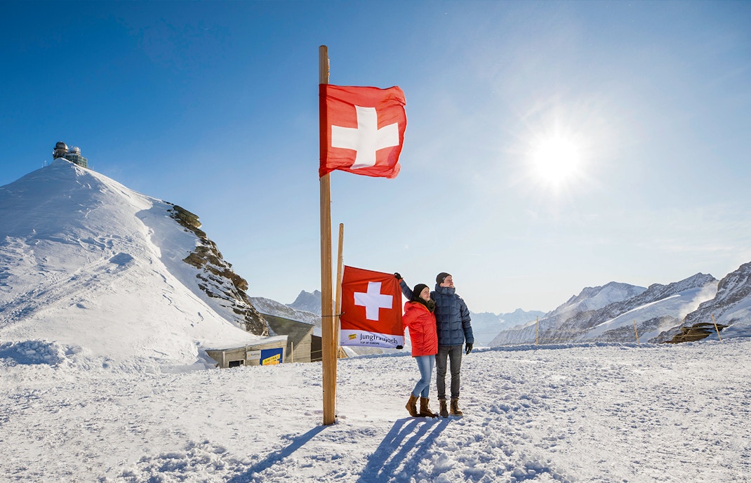 Visitors at Jungfraujoch Plateau with Swiss flags and snowy mountain backdrop.