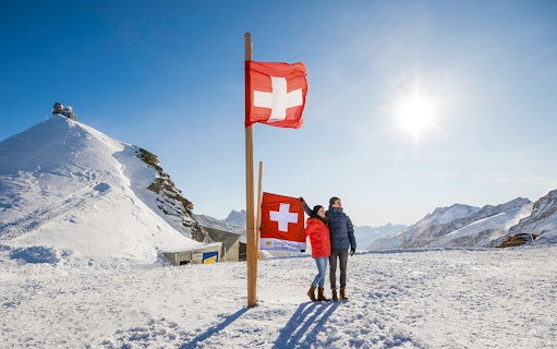 Visitors at Jungfraujoch Plateau with Swiss flags and snowy mountain backdrop.