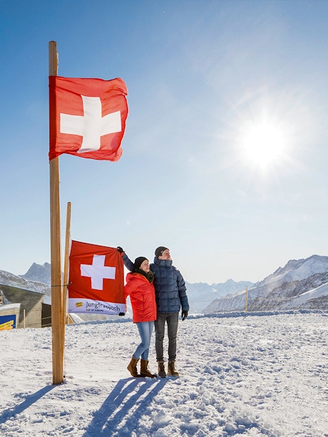 Visitors at Jungfraujoch Plateau with Swiss flags and snowy mountain backdrop.